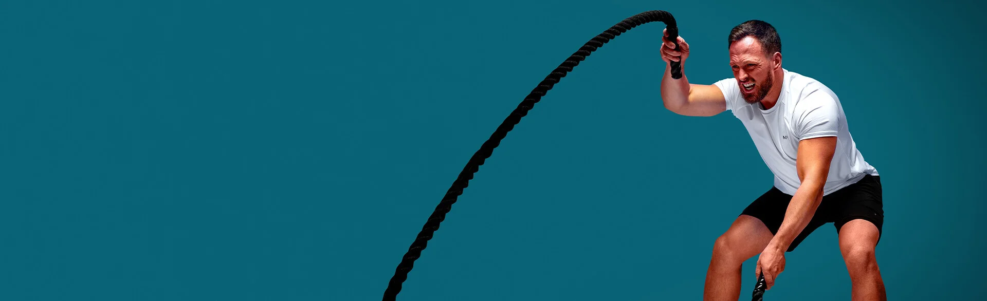 A man in a white t-shirt with the MP logo and black gym shorts working out with battle ropes on a light blue background.