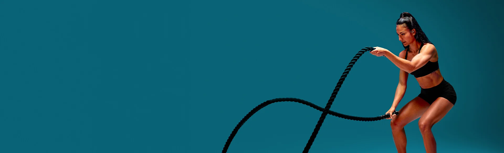 A woman wearing a black sports bra and black gym shorts and exercising using battle ropes.